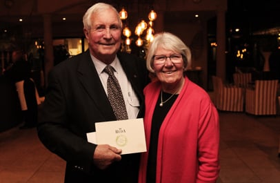Smiling elderly man and woman posing indoors, he holding a cream envelope with the Reda logo