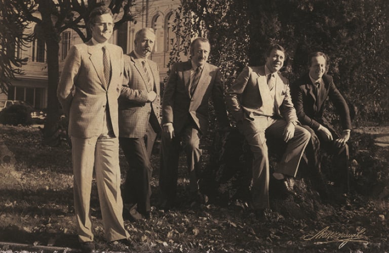 Five men in suits pose outdoors in a park in a vintage sepia photograph, with a building behind them