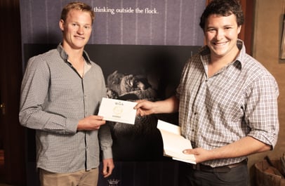 Two smiling men in checked shirts hand over a white envelope in front of a "thinking outside the flock" banner