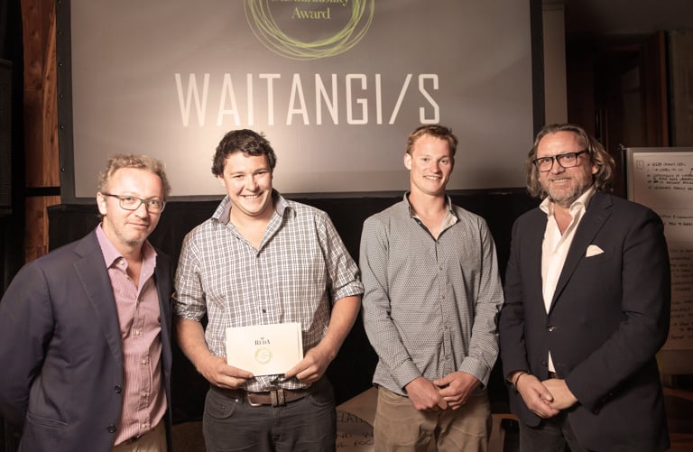 Four men pose at an award event, one holding a Reda envelope, in front of a screen reading "WAITANGI/S".