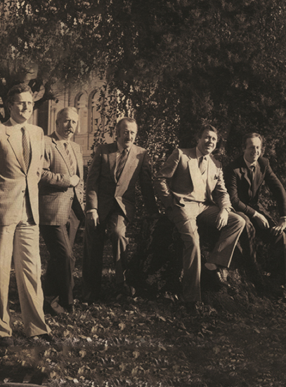 Five men in suits pose outdoors among trees and fallen leaves in a sepia vintage photo.