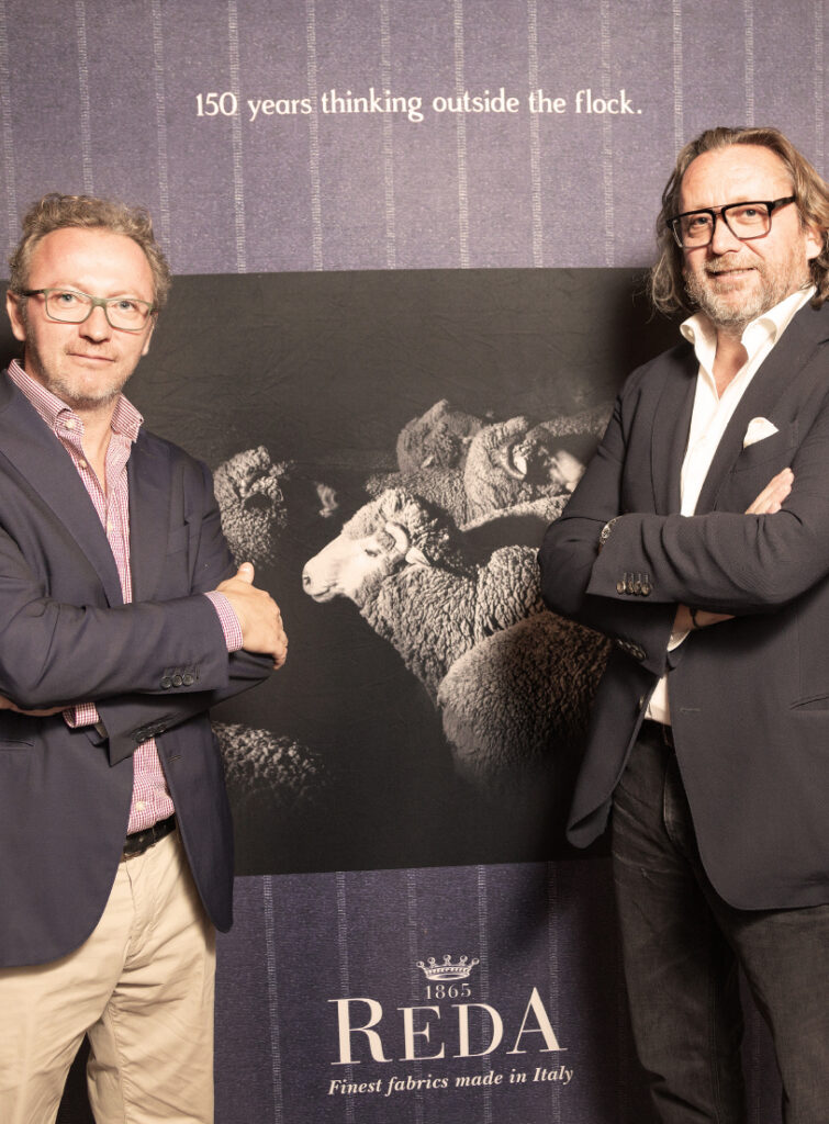 Two men with crossed arms pose before a Reda 1865 banner showing sheep: "150 years thinking outside the flock".