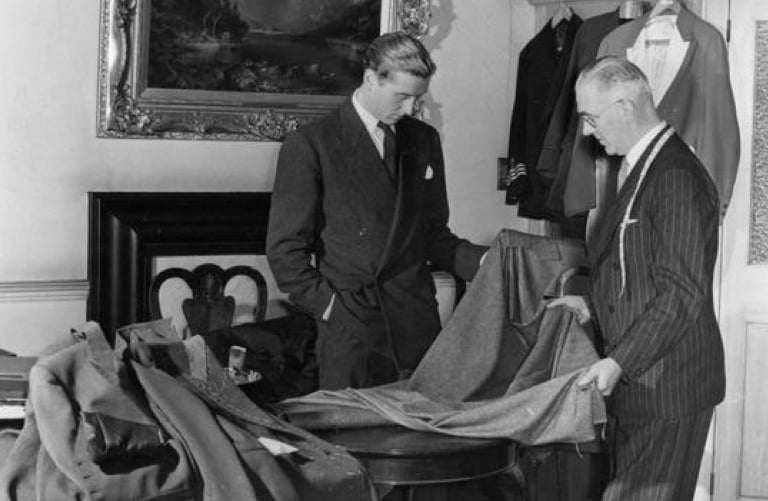 Two men in suits examine a heavy woolen fabric draped over a table in a room with a framed painting.