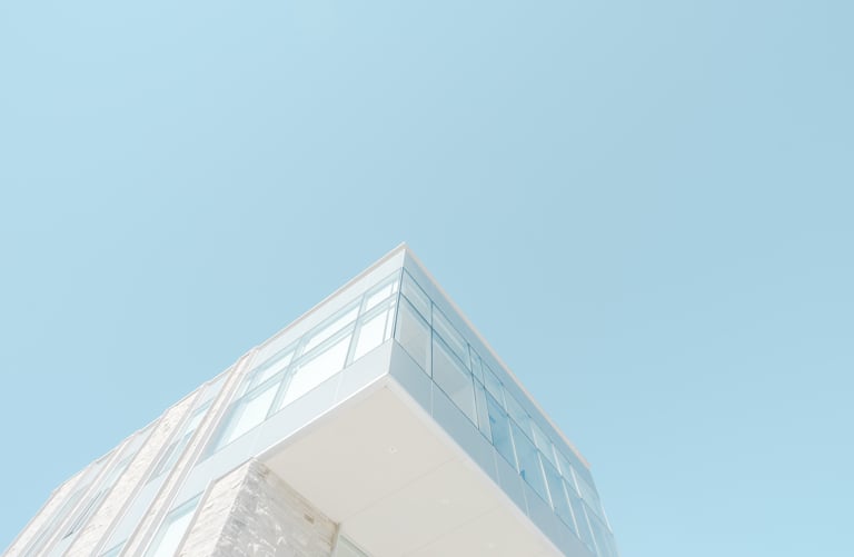 Corner of a modern white building with glass windows and stone cladding, seen from below against a clear blue sky
