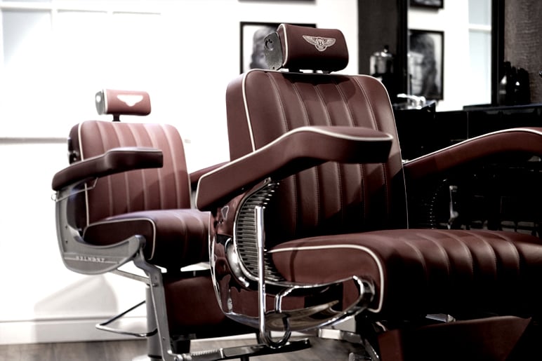Two burgundy leather barber chairs with cream piping and winged emblem on headrests in a barber shop