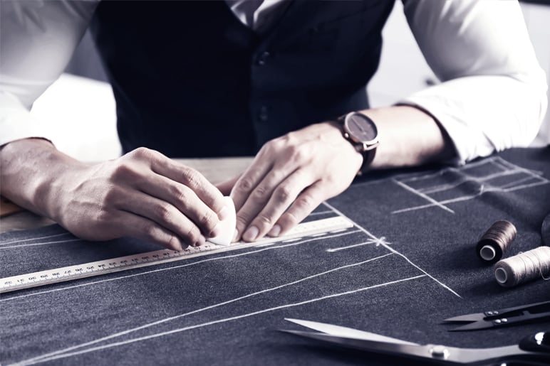 Tailor in a vest marking lines on dark grey wool fabric with chalk and a ruler, with thread spools and scissors nearby