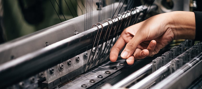 Mano che tocca i licci metallici e i fili di ordito all'interno di un telaio industriale per la tessitura
