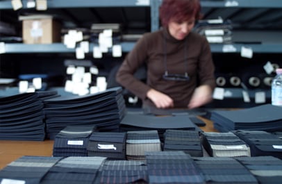 Woman in a brown sweater sorting dark fabric pieces in a workshop, with stacked swatches in the foreground