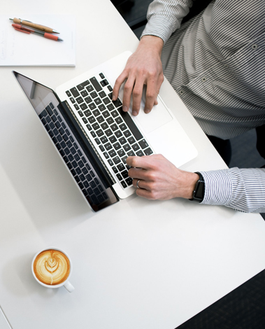 Man in a striped shirt typing on a laptop at a white desk, with a latte and pencils nearby