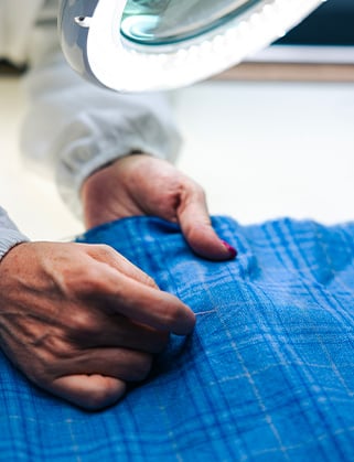 Hands sewing blue plaid fabric under a magnifying lamp with a ring of lights