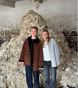 Two women smile in front of a large pile of raw wool in a building with whitewashed brick walls.