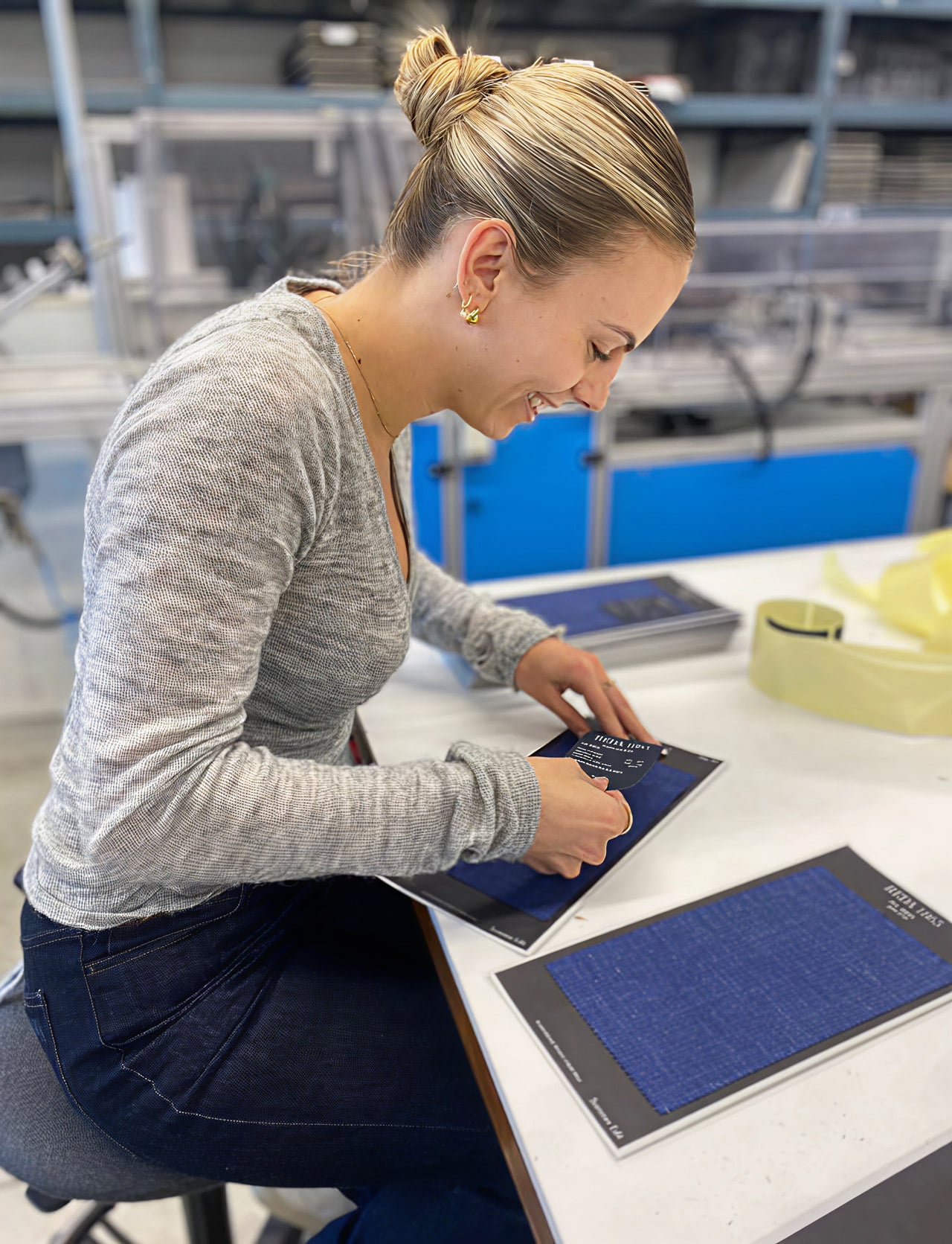 Smiling woman in a workshop attaches a label to a blue fabric swatch card on a white table