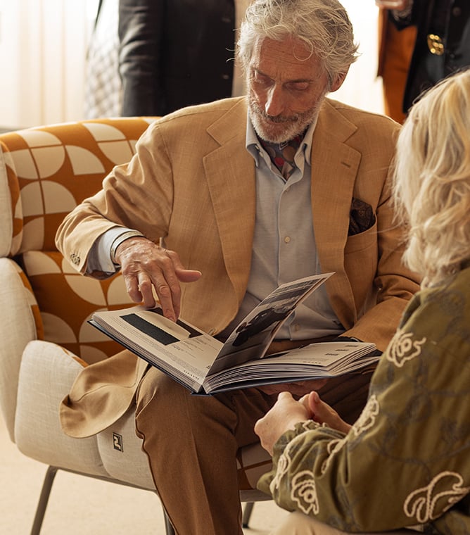 Elderly man in a beige jacket points at an open book while seated beside a blonde woman in an armchair.
