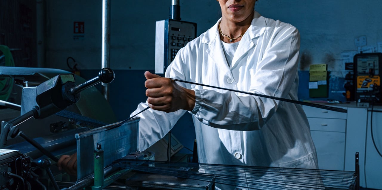 Worker in a white lab coat holds a taut thin strip of material beside industrial machinery in a workshop