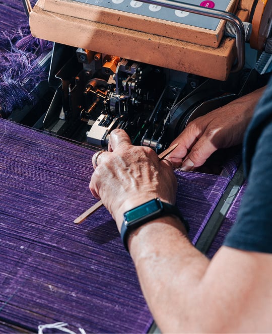 Hands guiding purple woven fabric with a wooden tool at a textile machine in a mill