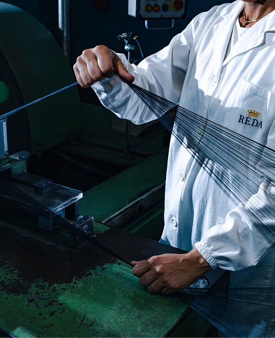 Worker in a white REDA coat holds taut fine yarn threads stretched across a green textile machine