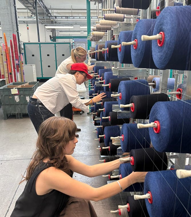 Three people handle blue and black yarn cones on a large spool rack in a textile mill.