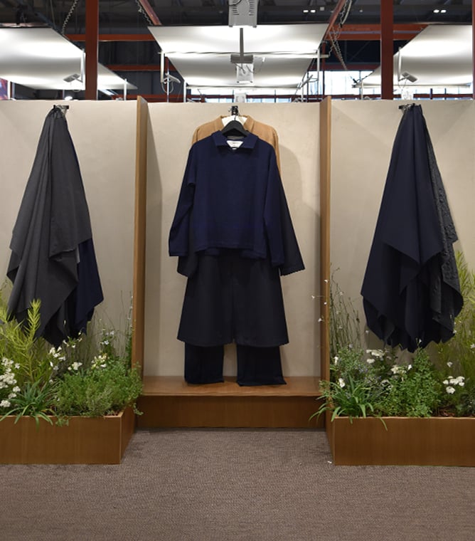 Navy top and dark trousers on a hanger between two draped dark capes, with plants in wooden planters