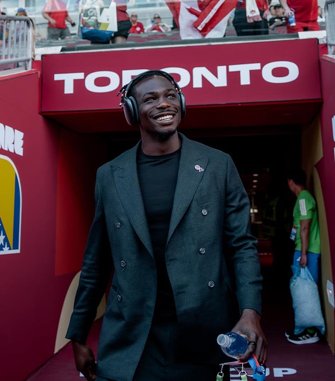 Smiling man in headphones and a dark green double-breasted jacket walks out of a tunnel marked "TORONTO"