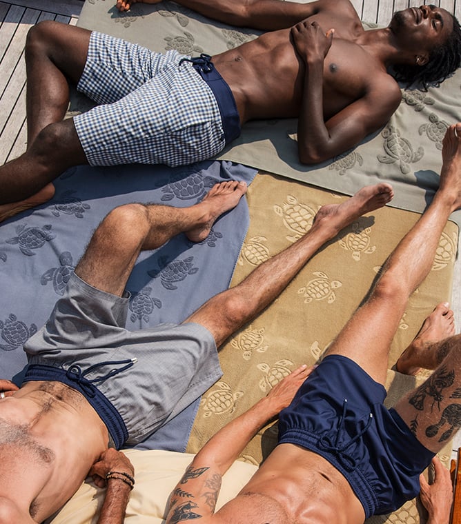Three men in swim shorts lie on turtle-patterned beach towels on a wooden deck