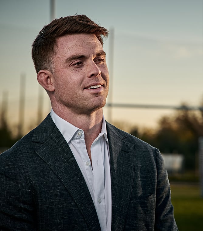 Smiling man in a dark textured wool jacket and white shirt, looking aside on a sports field at sunset