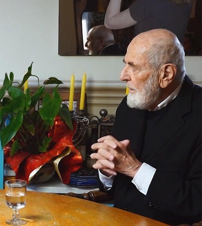 Elderly white-bearded man in a dark jacket sits at a wooden table, hands clasped, beside a plant and candles.