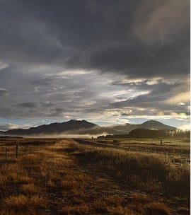 Frost-covered grassy pasture with a fence and dark mountains beneath a dramatic cloudy sky at sunrise