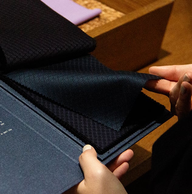 Hands leafing through a swatch book of dark navy textured wool fabrics with a small diamond pattern
