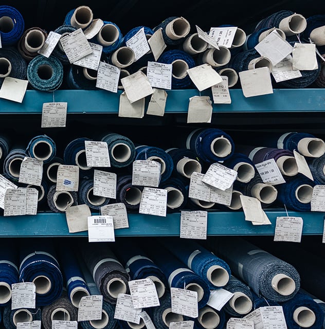Rolls of blue and grey fabric with paper labels stacked on teal warehouse shelves, tags reading Reda SPA