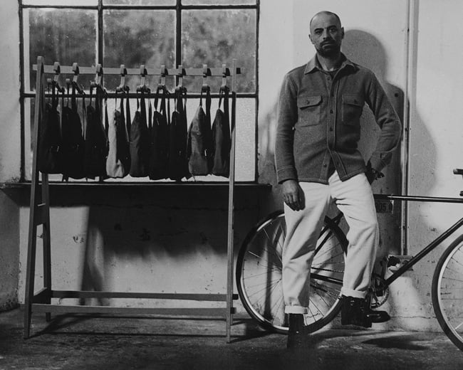 Uomo in camicia-giacca di lana e pantaloni chiari appoggiato a una bici accanto a un appendiabiti con gilet.