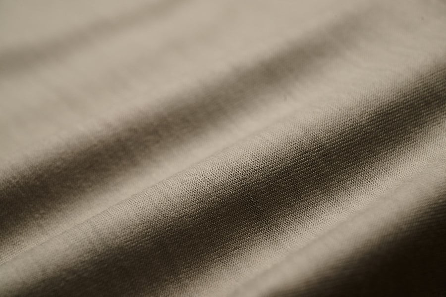 Close-up of soft beige-taupe jersey fabric with gentle folds and shadows, showing its fine knit texture