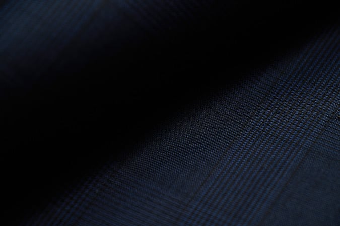 Close-up of dark navy wool fabric with a subtle glen check pattern, softly lit in the shadows