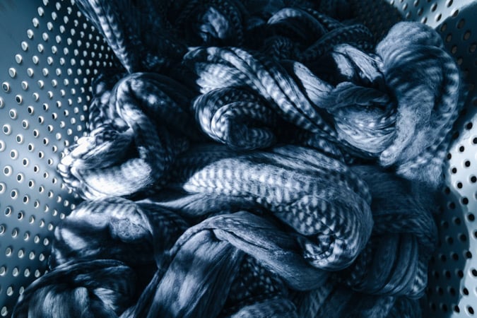 Blue-toned wool fibers twisted in loose coils inside a perforated metal colander, lit by striped shadows