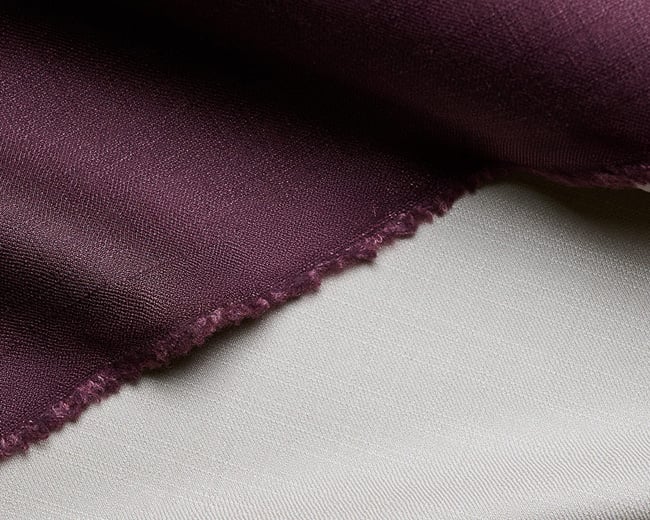 Close-up of plum purple wool fabric with a frayed edge, folded over a light grey fabric