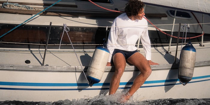 Man in white long-sleeve top and shorts sits on a sailboat's edge, feet in the water, beside two fenders