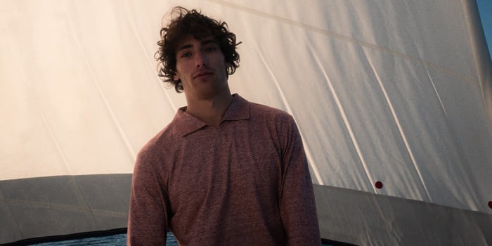 Young man with curly hair in a reddish knit polo shirt stands before a large white sail at sunset.