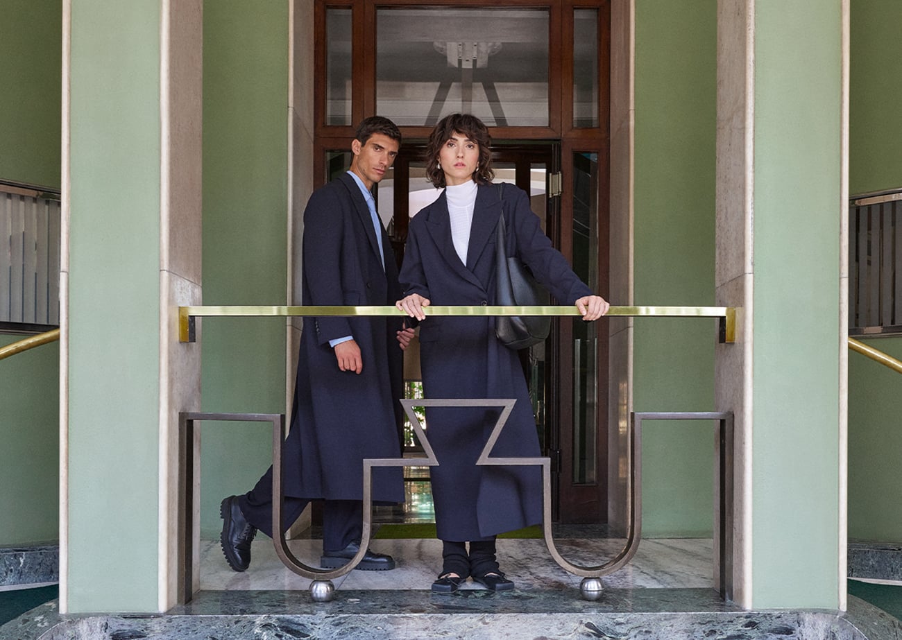 Man and woman in long navy coats stand behind a brass railing in a green-walled marble entrance