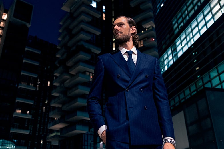 Man in a navy pinstripe double-breasted suit and tie stands at night among modern high-rise buildings