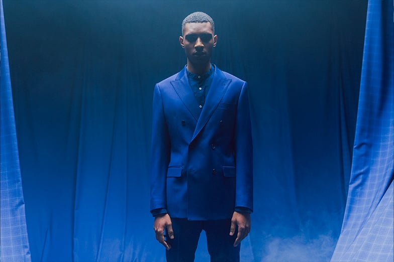 Man in a blue double-breasted suit stands before blue draped fabric, with checked curtains at the sides