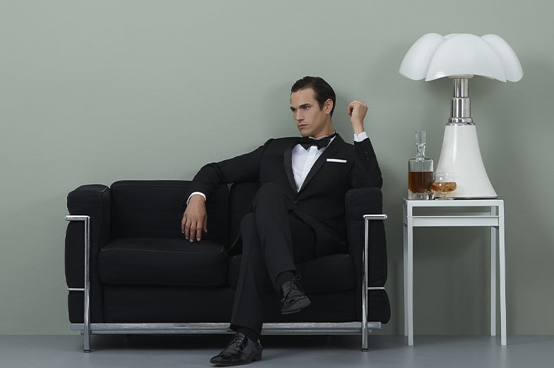 Man in a black tuxedo and bow tie sits on a black leather sofa beside a table with a lamp and whisky.