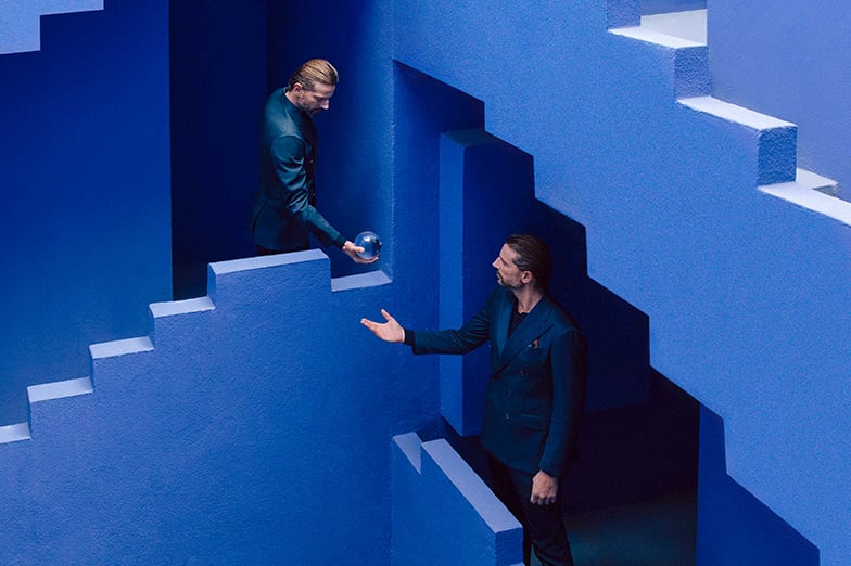 Two men in dark suits on blue stepped walls; one holds a glass sphere while the other reaches out.