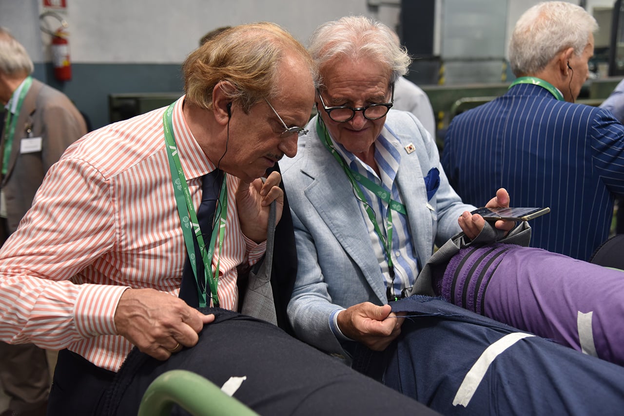 Two men with lanyards examine a piece of dark blue fabric together, one holding a phone, in a factory.