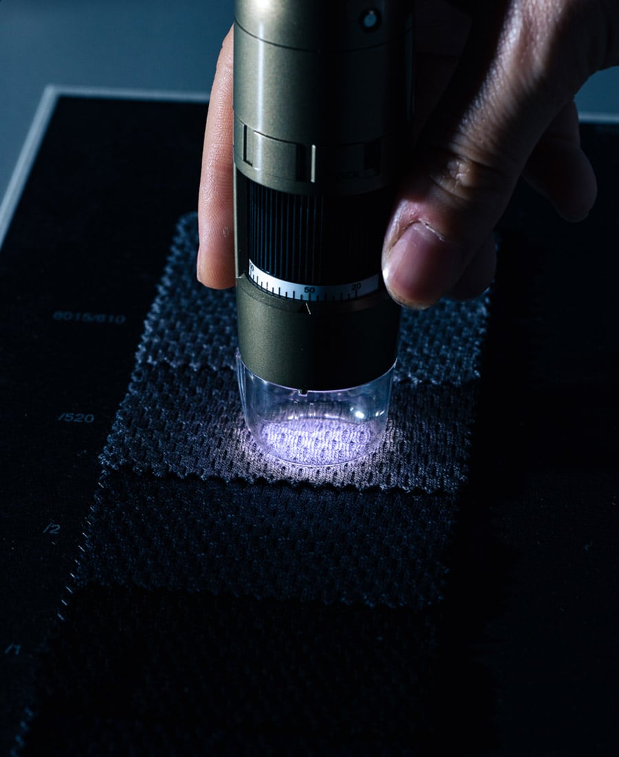 Hand holding a digital microscope over a dark knitted fabric swatch to inspect its texture