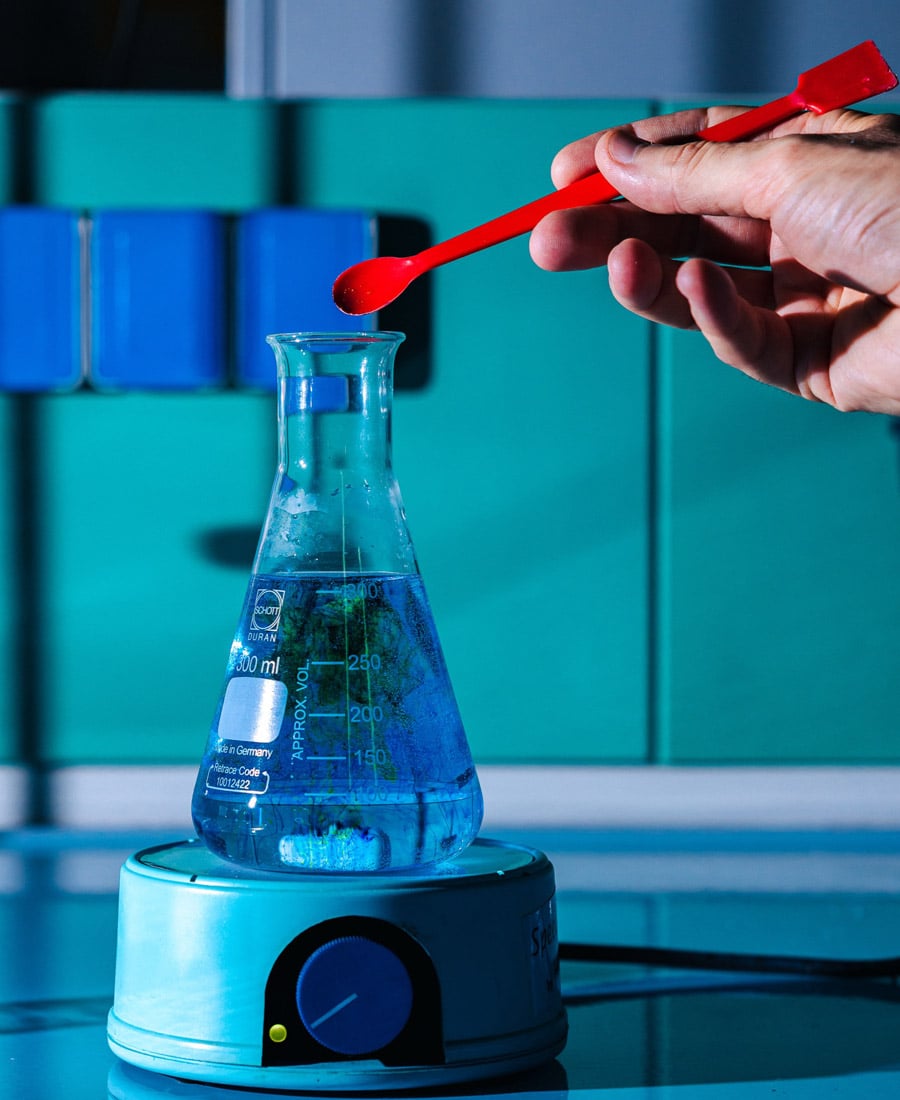 Hand holding a red spatula over a 300 ml Erlenmeyer flask of blue liquid on a magnetic stirrer in a lab