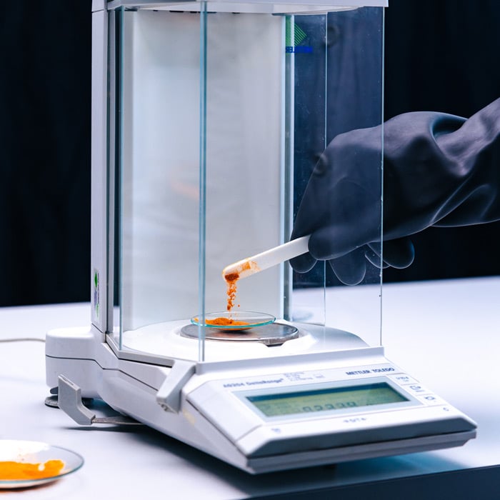 Gloved hand pours orange powder from a spatula onto a watch glass on a Mettler Toledo analytical balance