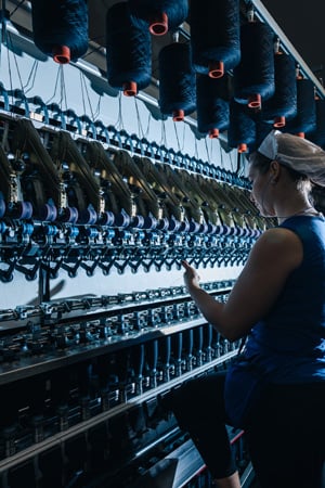 Woman in a hairnet tends a textile machine with rows of dark thread spools in a spinning mill.