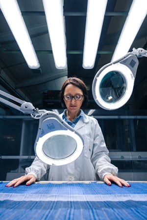 Woman in a white lab coat inspects a blue woven fabric under magnifying lamps and bright overhead lights.