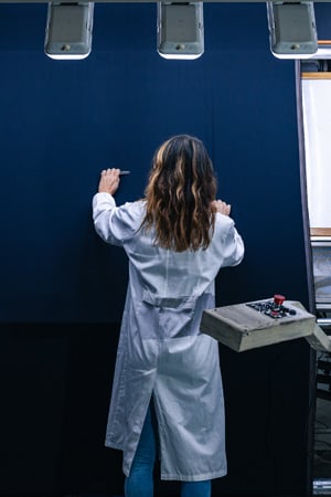 Woman in a white lab coat seen from behind, inspecting a large dark blue fabric panel beside a control console