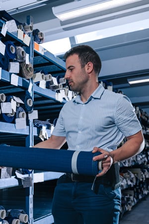 Man in a striped shirt holds a rolled blue fabric beside warehouse shelves stocked with fabric rolls.