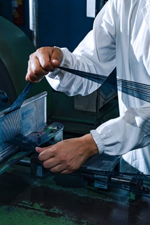 Worker in a white coat guides fine dark threads through a comb guide on a textile machine